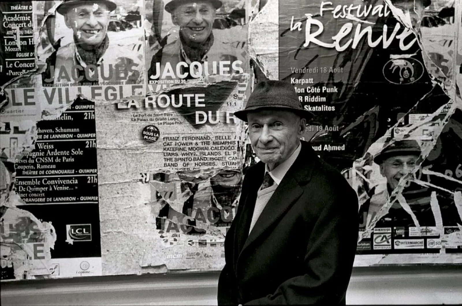 Jacques Villeglé en chapeau devant un mur d'affiches lacérées à son nom, portrait noir et blanc