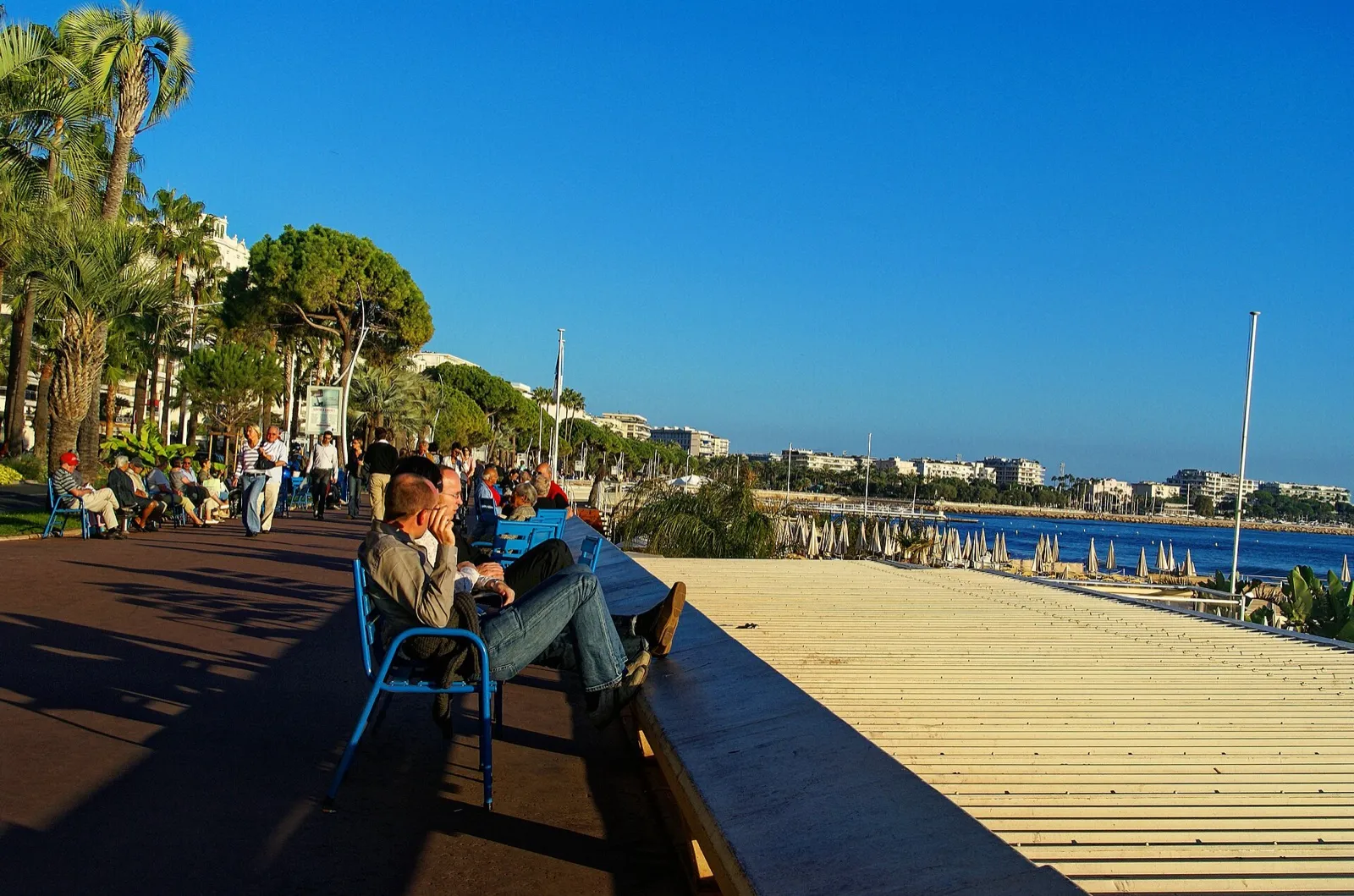 Promeneurs sur les chaises bleues de la promenade de la Croisette face à la baie de Cannes
