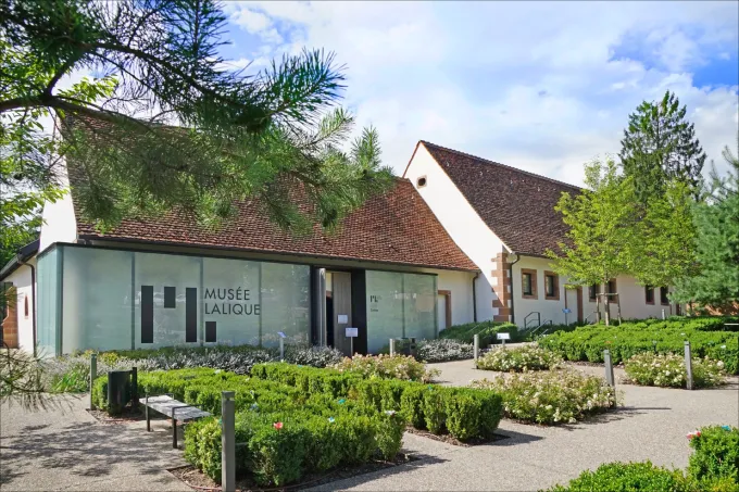 Bâtiment vitré du musée Lalique à Wingen-sur-Moder, dans son jardin de buis taillés, sous un ciel bleu