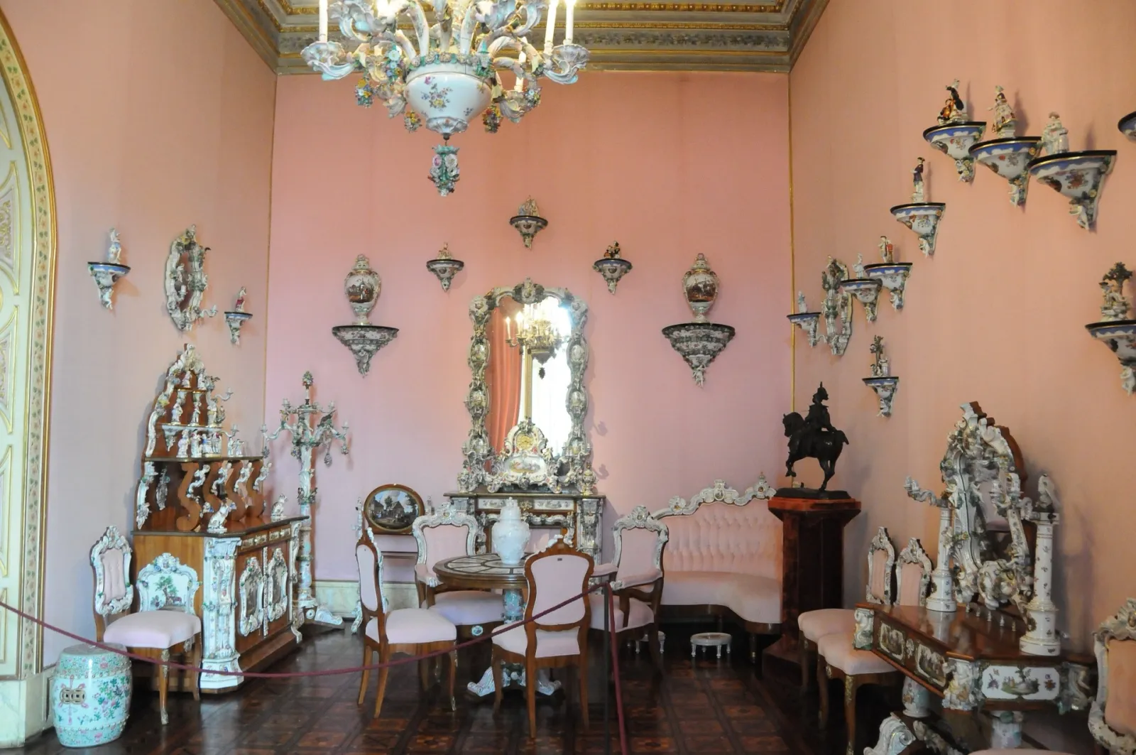Salon aux murs rose poudré orné de porcelaines, d'un grand lustre à pampilles et de mobilier ancien