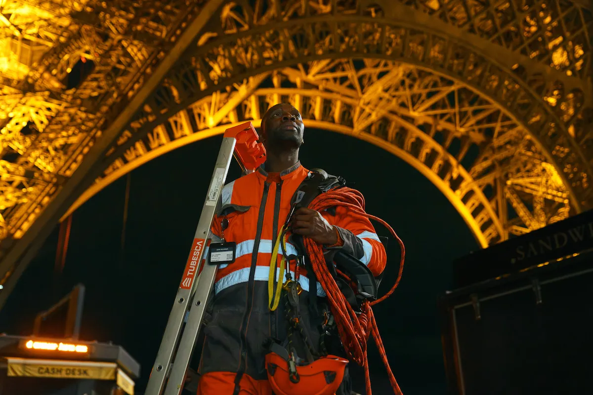 Assane Diop en tenue de technicien, échelle et câbles à l'épaule, sous les arches illuminées de la tour Eiffel, Lupin partie 4
