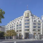 Façade de la Maison Louis Vuitton sur les Champs-Élysées, à Paris, sous un ciel bleu, malle monogramme à droite