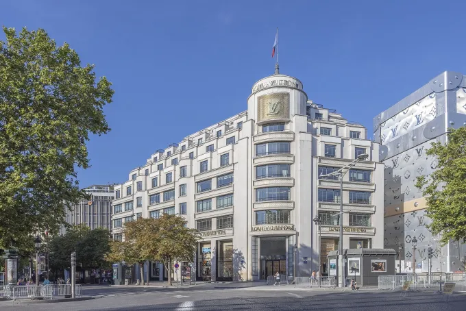 Façade de la Maison Louis Vuitton sur les Champs-Élysées, à Paris, sous un ciel bleu, malle monogramme à droite