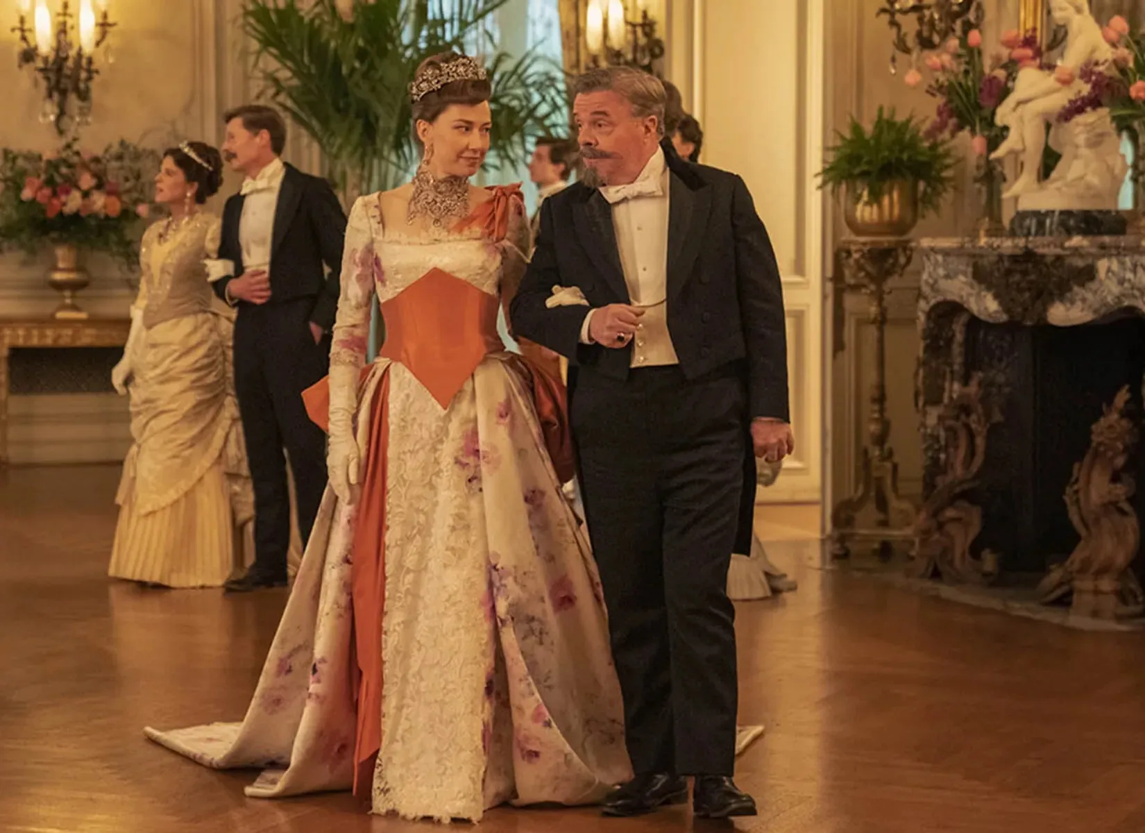 Carrie Coon en Bertha Russell, diadème et collier de pierres, au bras de Nathan Lane dans la salle de bal de The Elms